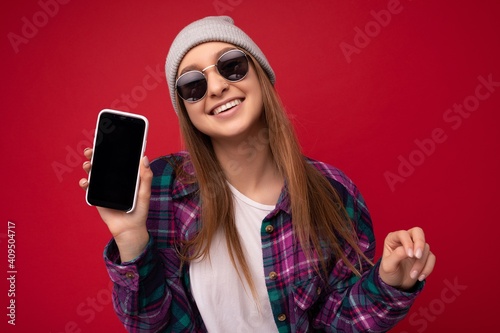 Closeup photo of attractive smiling positive young blonde woman wearing stylish purple shirt and casual white t-shirt grey hat and sunglasses isolated over red background holding in hand and showing