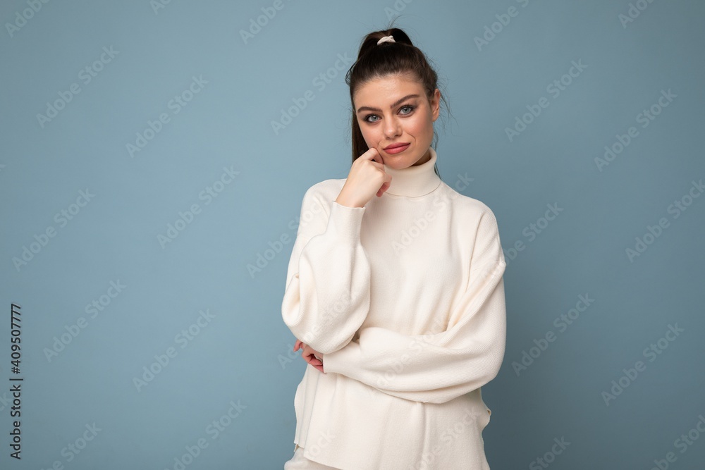 Young sad brunette woman with wearing white casual sweater isolated over blue background looking confident at the camera with crossed arms and hand raised on chin Young sad brunette woman with wearing white casual sweater isolated over blue background looking confident at the camera with crossed arms and hand raised on chin