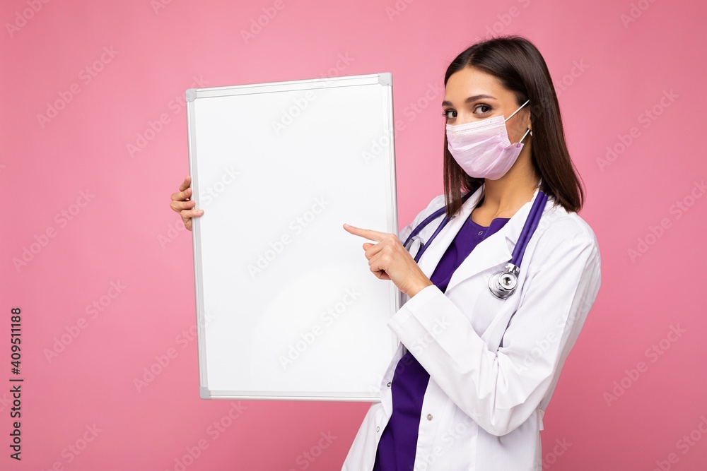 Beautiful brunette female nurse in protective face mask and white medical coat holding an empty magnetic board isolated on pink background Beautiful brunette female nurse in protective face mask and white medical coat holding an empty magnetic board isolated on pink background