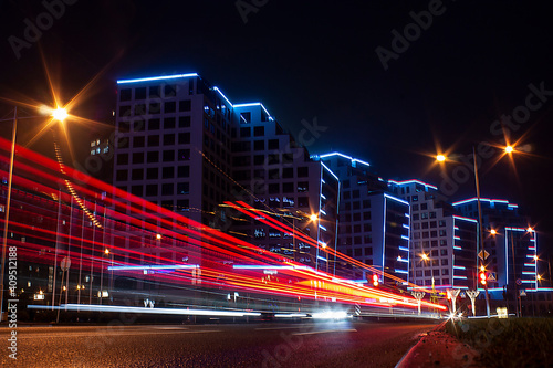 Horizontal long exposure photo with night city street  modern buildings along the road with blue illumination and red tail light trails of vehicle