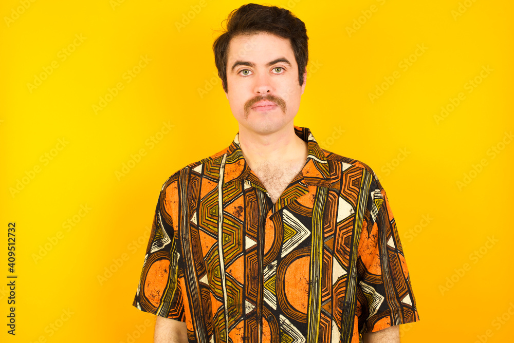 Joyful young Caucasian man wearing printed shirt against yellow wall looking to the camera, thinking about something. Both arms down, neutral facial expression. Joyful young Caucasian man wearing printed shirt against yellow wall looking to the camera, thinking about something. Both arms down, neutral facial expression.
