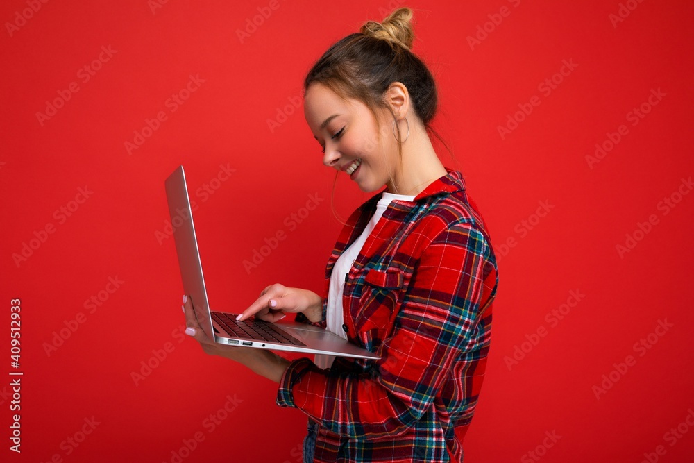 side profile photo of pretty smiling young woman holding netbook looking at screen typing on keyboard wearing red shirt isolated over red wall background side profile photo of pretty smiling young woman holding netbook looking at screen typing on keyboard wearing red shirt isolated over red wall background