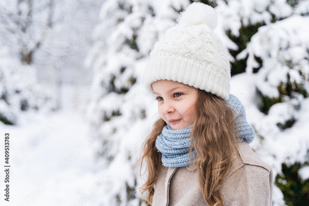 Little beautiful stylish girl in winter hat, woolen coat, blue scarf, snood with long hair. Kid walking, playing in forest, park among trees covered with snow. Country house yard. Fashionable image Little beautiful stylish girl in winter hat, woolen coat, blue scarf, snood with long hair. Kid walking, playing in forest, park among trees covered with snow. Country house yard. Fashionable image