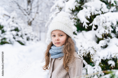 Little beautiful stylish girl in winter hat, woolen coat, blue scarf, snood with long hair. Kid walking, playing in forest, park among trees covered with snow. Country house yard. Fashionable image