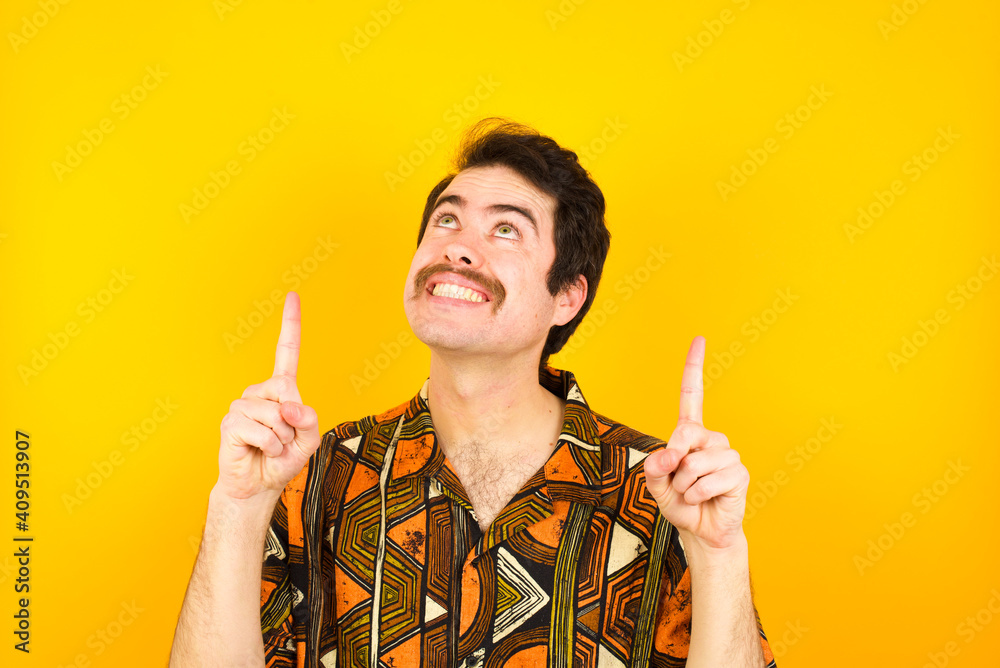 Successful friendly looking young Caucasian man wearing printed shirt against yellow wall exclaiming excitedly, pointing both index fingers up, indicating something. Successful friendly looking young Caucasian man wearing printed shirt against yellow wall exclaiming excitedly, pointing both index fingers up, indicating something.