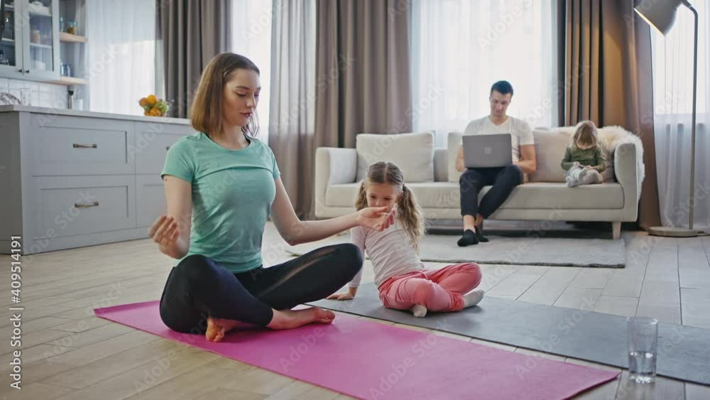Mother teaching her little daughter to meditate, sitting together in lotus positions, father and son on background Mother teaching her little daughter to meditate, sitting together in lotus positions, father and son on background
