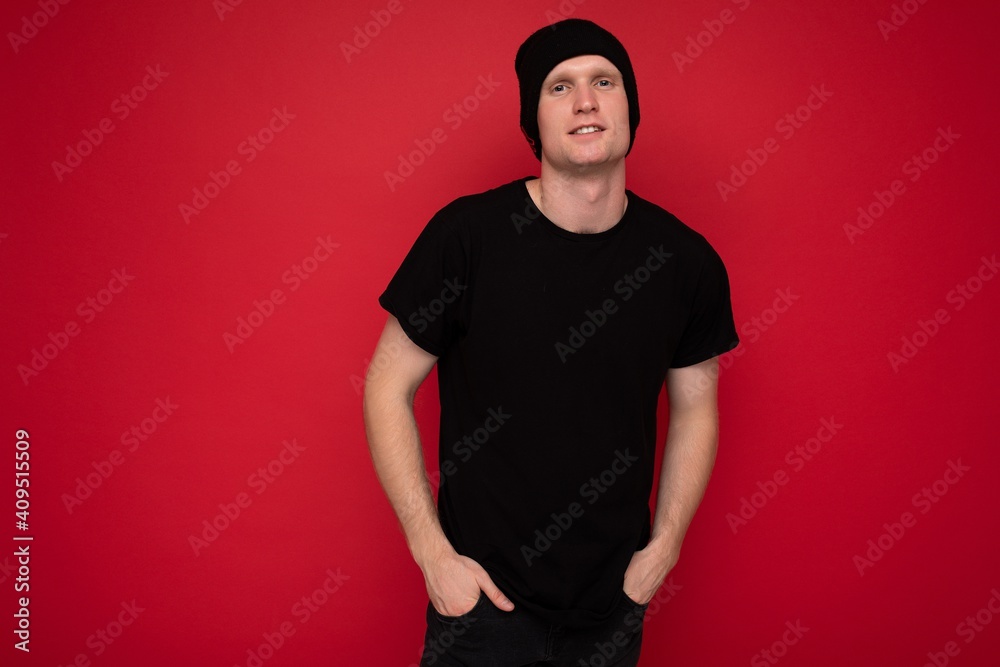 Shot of handsome happy smiling young man standing isolated over red background wall wearing black t-shirt for mockup and black hat and looking at camera Shot of handsome happy smiling young man standing isolated over red background wall wearing black t-shirt for mockup and black hat and looking at camera