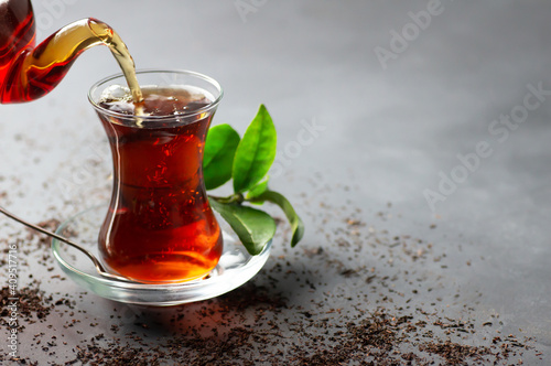 Fototapet Glass cup of black tea pouring from teapot with fresh tea leaves, traditional tu