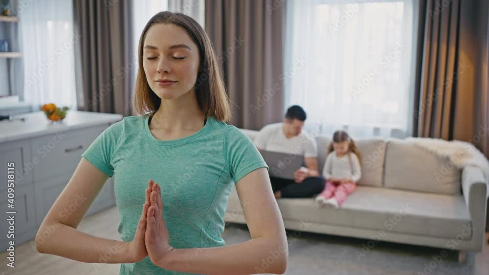 Close up shot of balanced woman practicing breathing exercise at home, her husband and daughter playing on laptop Close up shot of balanced woman practicing breathing exercise at home, her husband and daughter playing on laptop