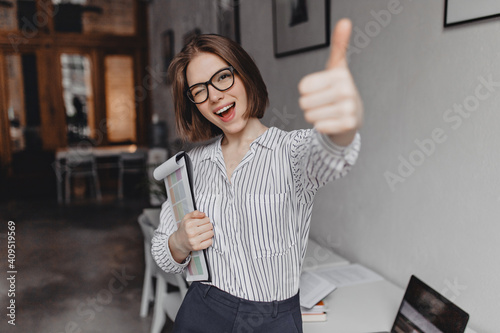 Young woman in office style clothes and glasses holds tablet with documents, winks and shows thumb up