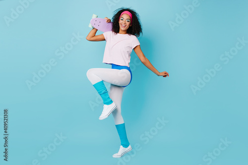 Fototapeta Naklejka Na Ścianę i Meble -  Studio shot of jumping african girl in aerobics form. Graceful black young woman posing with skateboard on blue background.