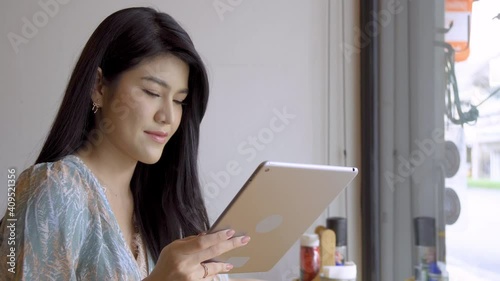 Beautiful young Asian woman sitting at counter and drinking beverage in coffee shop, using digital tablet with happy mood and smiling.