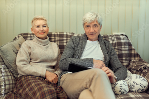 Attractive middle aged female sitting comfortably on sofa with her gray haired husband, relaxing together on cozy evening, surfing Internet on mobile phone. People, aging, marriage and domestic life