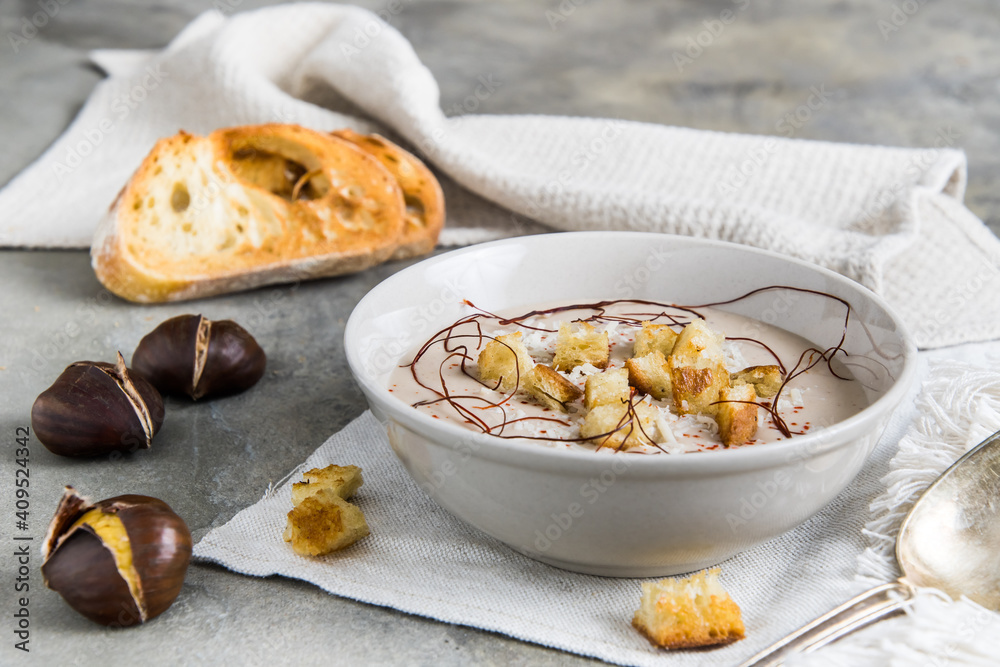 Chestnut soup with parmesan, croutons and chilli flakes and threads on grey concrete background, decorated with roasted bread and chestnuts Chestnut soup with parmesan, croutons and chilli flakes and threads on grey concrete background, decorated with roasted bread and chestnuts