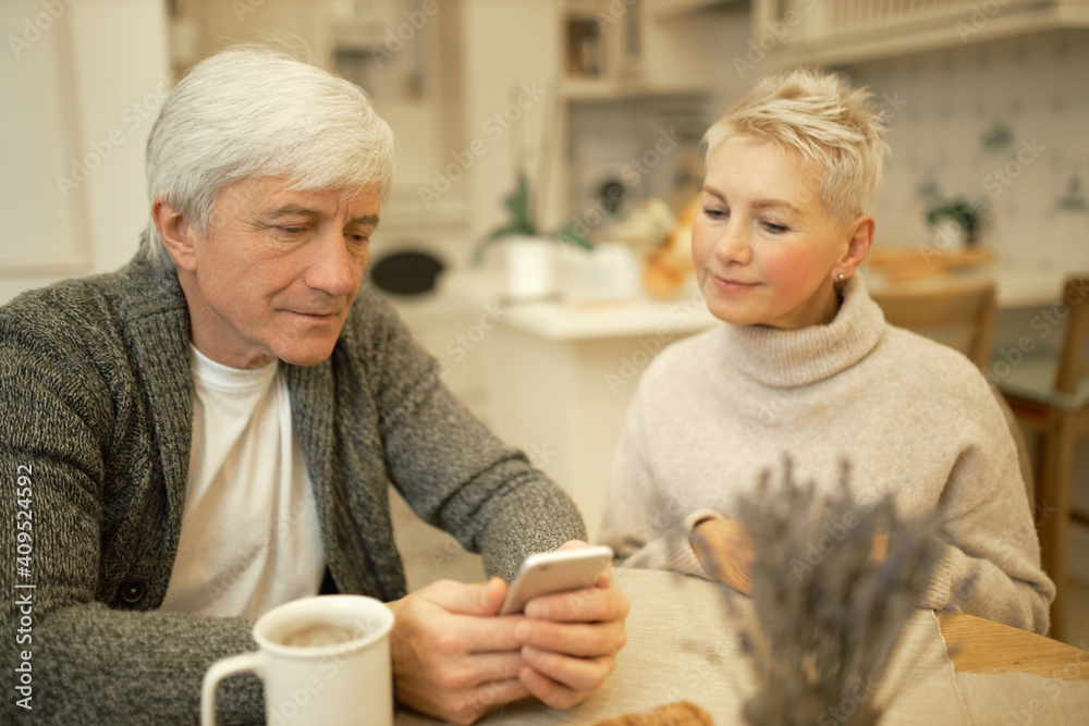 Serious elderly man with gray hair and wrinkles typing text message on cell phone, drinking tea in kitchen with wife. Cute senior couple relaxing indoors, using electronic gadget during breakfast Serious elderly man with gray hair and wrinkles typing text message on cell phone, drinking tea in kitchen with wife. Cute senior couple relaxing indoors, using electronic gadget during breakfast