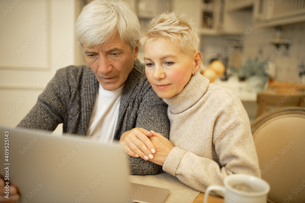 Serious elderly man and his blonde wife sitting in kitchen in front of open laptop, surfing internet together, signing up on website or social networks, looking at screen with concentrated expression Serious elderly man and his blonde wife sitting in kitchen in front of open laptop, surfing internet together, signing up on website or social networks, looking at screen with concentrated expression