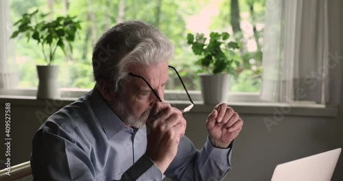 Head shot focused middle aged old man taking off glasses, making pause in computer work, suffering from dry eyes syndrome or relieving eyes strain, continue working online using modern tech gadget.