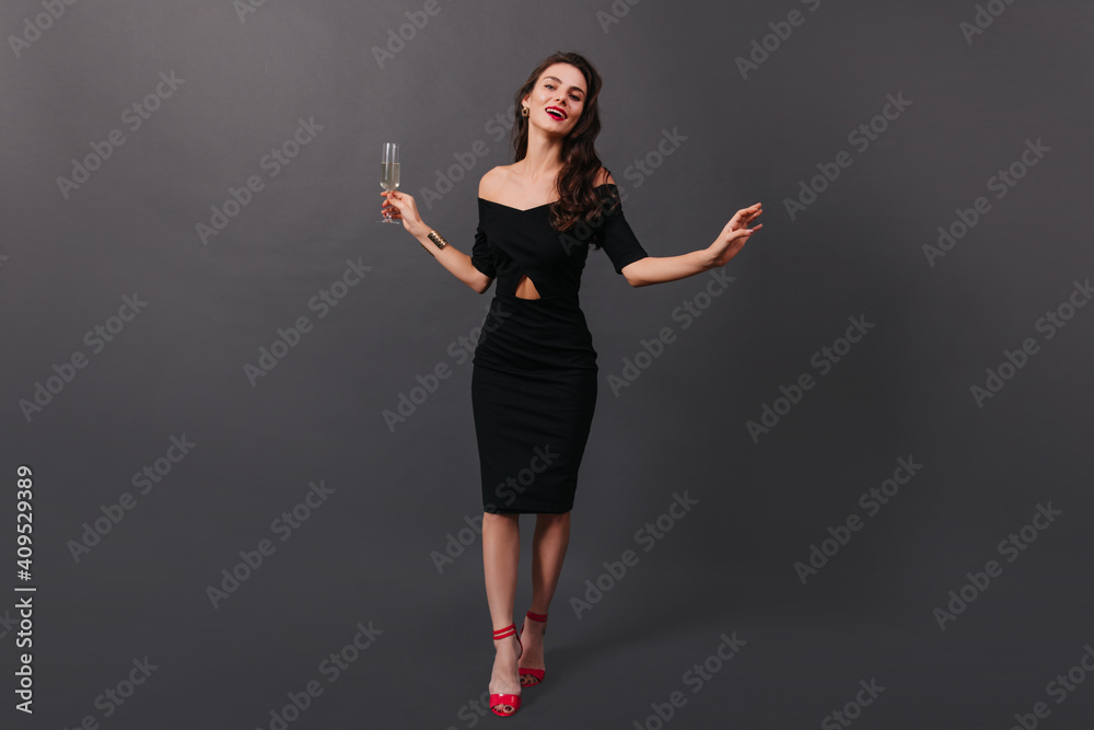 Full-length photo of woman in black fitting dress and in high heels posing on black background with glass of champagne in her hands