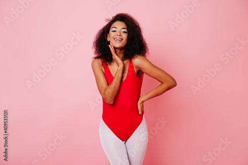 Lovely young woman in red bodysuit laughing on pink background. Studio shot of stylish african fitness girl.