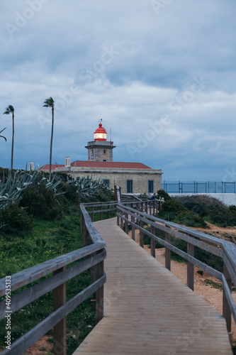 Lonely building of a lighthouse on cloudy storm day, wind bends palm trees. Wooden boardwalk leads to lighthouse in the distance. Moody cinematic photo