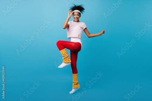 Fototapeta Naklejka Na Ścianę i Meble -  Inspired black woman enjoying aerobics. Full length view of glamorous african girl jumping on blue background.
