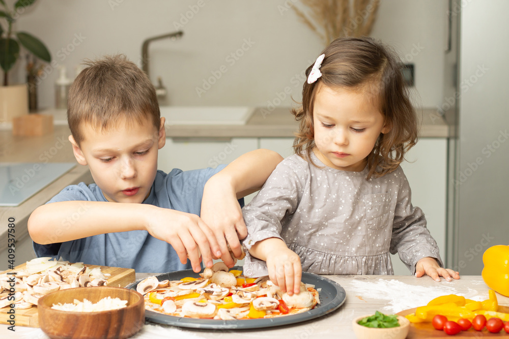 Cute little girl 2-4 in gray dress and boy 7-10 in T-shirt cooking pizza together in kitchen. Brother and sister cooking