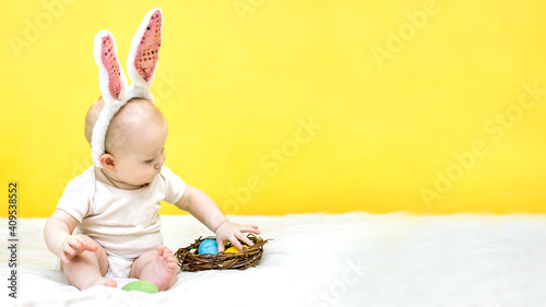 banner with child sitter with Easter bunny ears decorated with sequins sits near yellow wall. baby found colorful egg in nest. Easter hunt concept photo