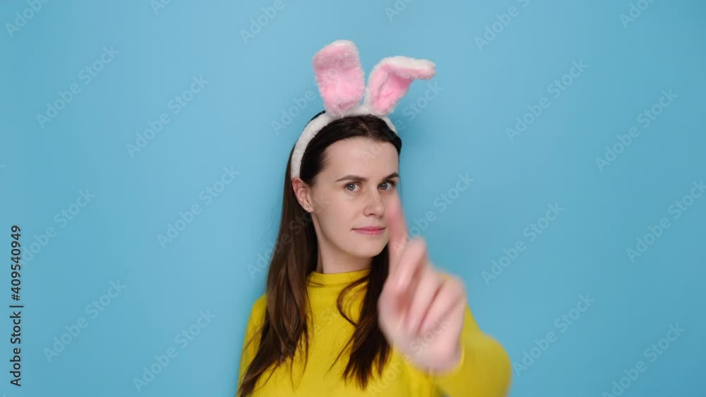 Serious young brunette woman with pink bunny fluffy ears is shaking her finger negatively, dressed in yellow sweater, models over blue studio background. Easter holiday and disapproval sign concept