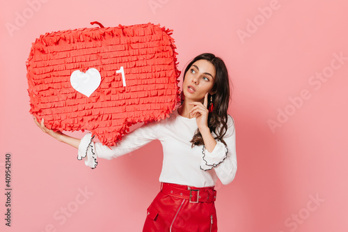 Long-haired brunette thoughtfully looks at huge red like from Instagram. Close-up shot of cute tanned girl photo