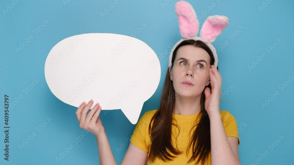 Pensive young woman pink bunny fluffy ears holding and pointing at paper blank speech bubble, wears yellow t-shirt, isolated on blue background studio with copy space. Easter holiday concept Pensive young woman pink bunny fluffy ears holding and pointing at paper blank speech bubble, wears yellow t-shirt, isolated on blue background studio with copy space. Easter holiday concept