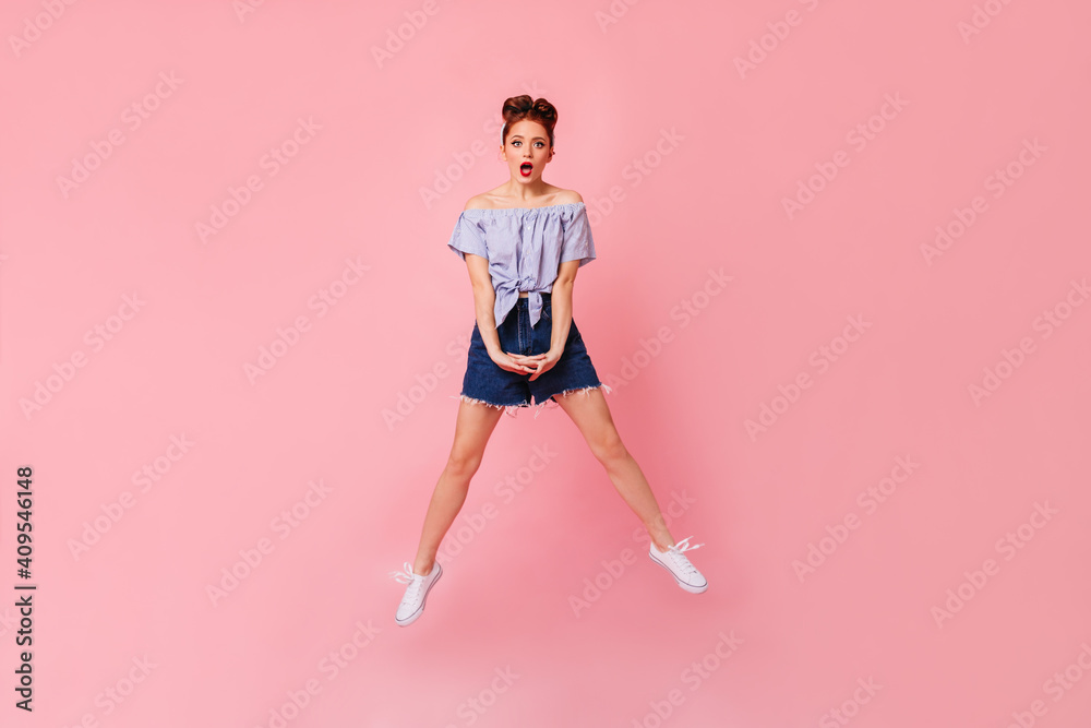Full length view of shocked woman in denim shorts and blouse. Studio shot of amazed pinup girl jumping on pink background.