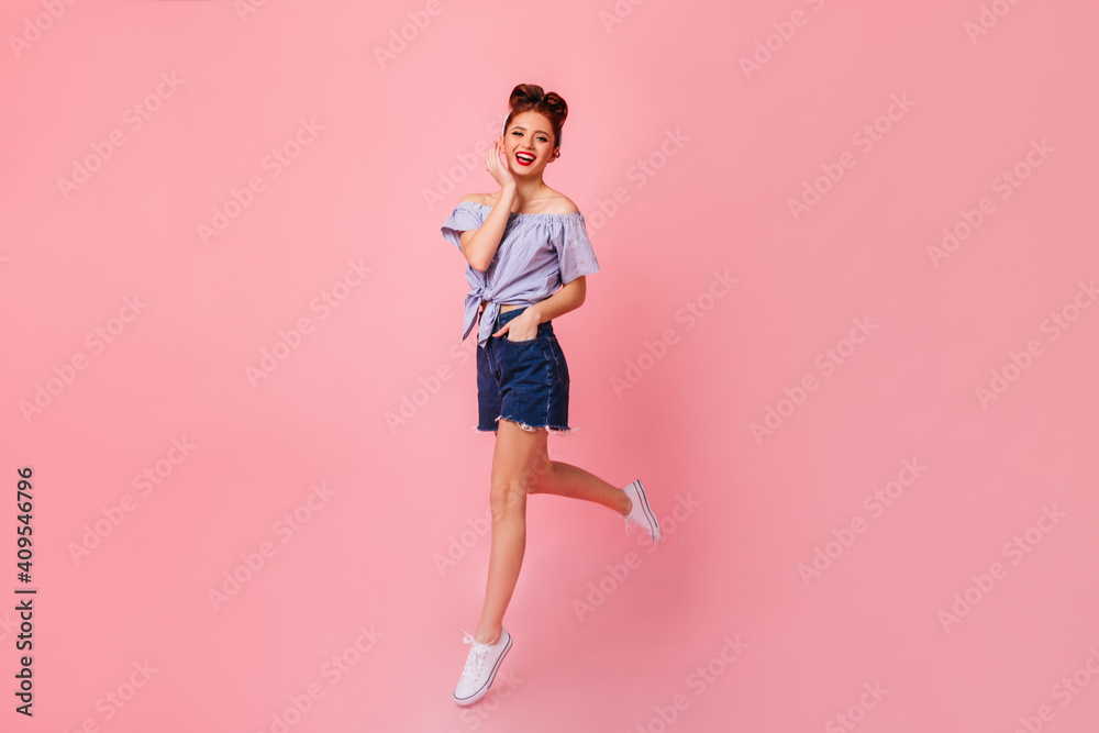 Laughing pinup girl posing with hand in pocket. Full length view of pretty ginger woman in denim shorts jumping on pink background. Laughing pinup girl posing with hand in pocket. Full length view of pretty ginger woman in denim shorts jumping on pink background.