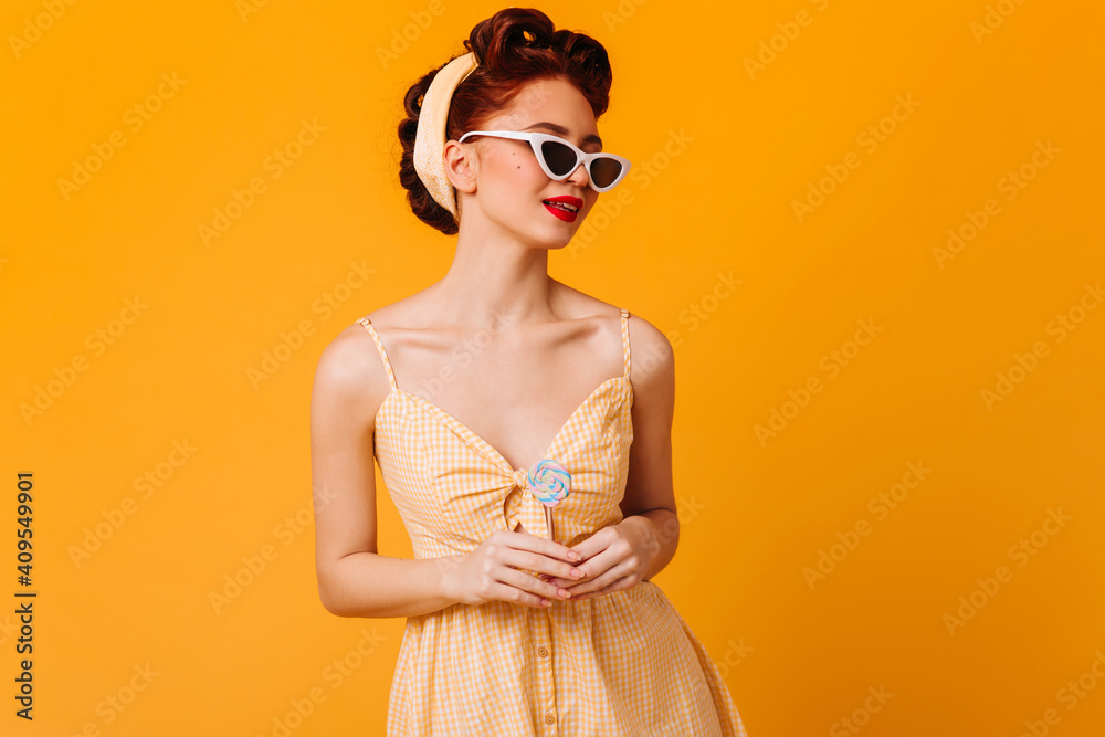 Adorable pinup woman posing in sunglasses. Studio shot of ginger girl with lollipop isolated on yellow background. Adorable pinup woman posing in sunglasses. Studio shot of ginger girl with lollipop isolated on yellow background.