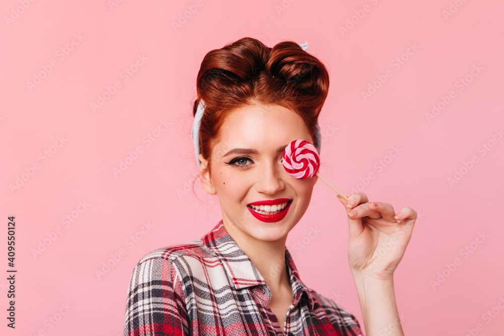 Smiling pinup girl eating red lollipop. Front view of woman in checkered shirt isolated on pink background. Smiling pinup girl eating red lollipop. Front view of woman in checkered shirt isolated on pink background.