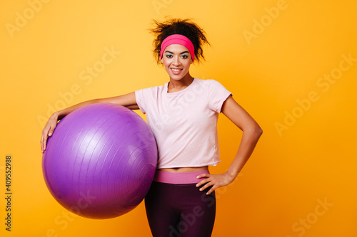 Cheerful african girl smiling after aerobics training. Studio shot of carefree young woman with fitness ball standing on yellow background. © Look!