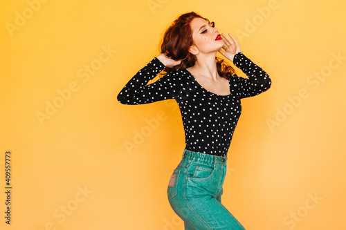 Joyful woman in green pants playing with curly hair. Studio shot of positive ginger girl dancing on yellow background.