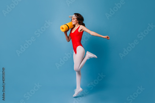 Fototapeta Naklejka Na Ścianę i Meble -  Healthy strong young girl of 80s doing sports on blue background. Woman in bodysuit and tights jumping with mat for fitness