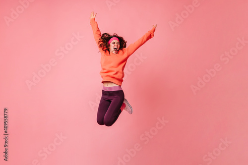 Active girl in orange hoodie and dark leggings vigorously jumping on pink background