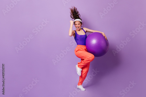 Woman in sports cap and bright top rejoices victory. Portrait of girl in red pants with fitball on purple background