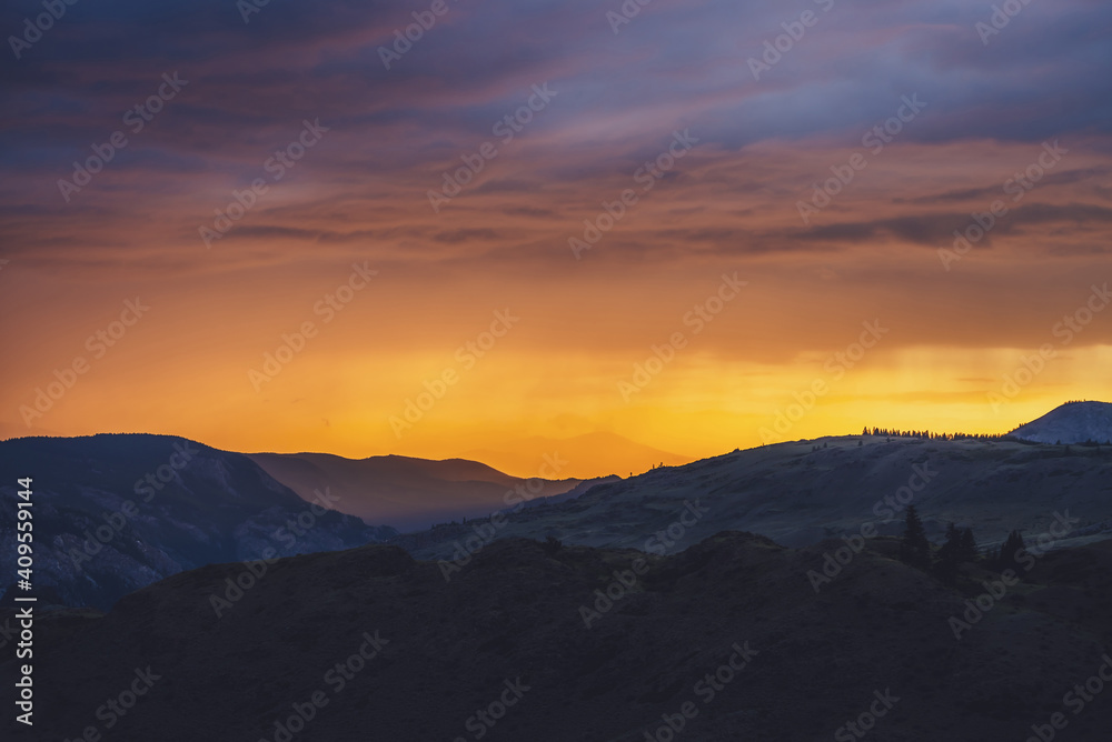 Atmospheric landscape with silhouettes of mountains with trees on background of vivid orange blue violet dawn sky. Colorful nature scenery with sunset or sunrise of illuminating color. Sundown paysage