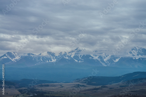 Scenic mountain landscape with great snowy mountain range among low clouds and green forest in valley at early morning. Atmospheric alpine scenery with blue white high mountain ridge under cloudy sky.