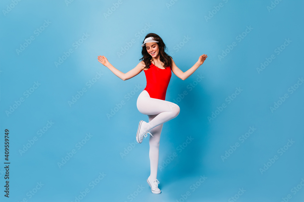 Obraz premium Girl from 80s jumps on blue background. Full-length shot of woman in fitness bodysuit and white leggings