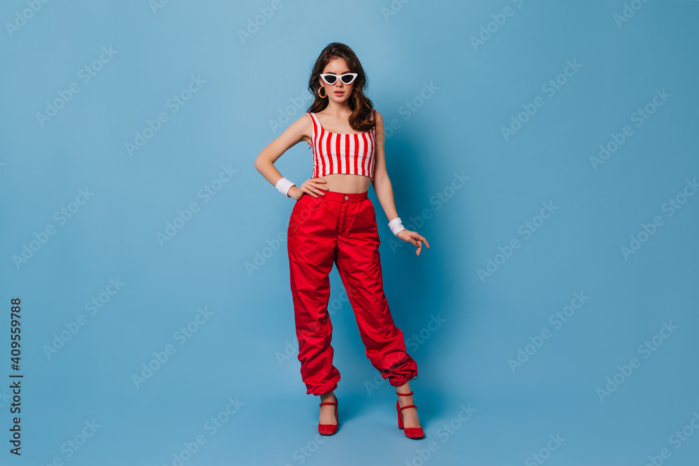 Stylish 80s girl in red pants and striped top poses on blue background. Full-length shot of brunette in white-rimmed sunglasses