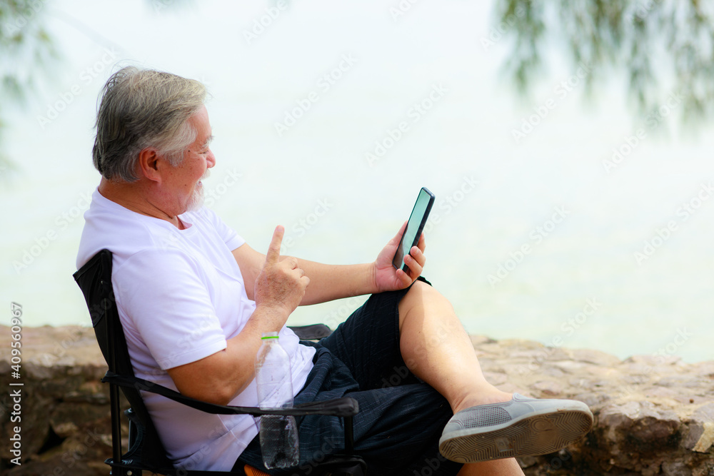 Asian senior male using mobile phone for video call communication with his family while vacation next to the beach.
