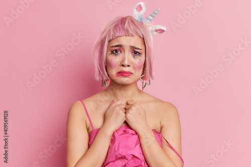 Doleful upset crying woman has spoiled makeup looks stressfully at camera has pink bob hair wears dress poses against pink background frowns face isolated over rosy background. Negative emotions