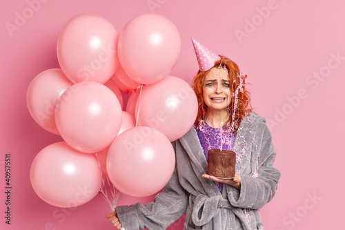 Dissatisfied redhead European woman cries from despair celebrates birthday alone holds cake with burning candle wears party hat and dressing gown holds balloons isolated over pink background photo