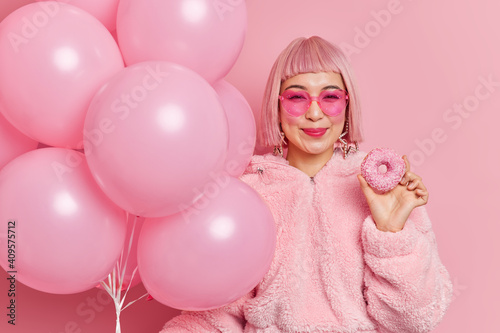 Happy pink hared girl in heart shaped sunglasses and fur coat holds delicious sweet doughnut bunch of inflated balloons smiles positively poses against rosy background comes on festive event © wayhome.studio 