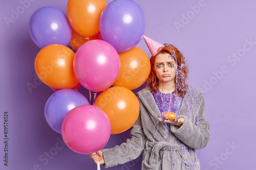 Waist up shot of unhappy ginger woman frowns face and looks dissatisfied at camera has bad mood after birthday celebration holds cupcake with burning candle and inflated multicolored balloons