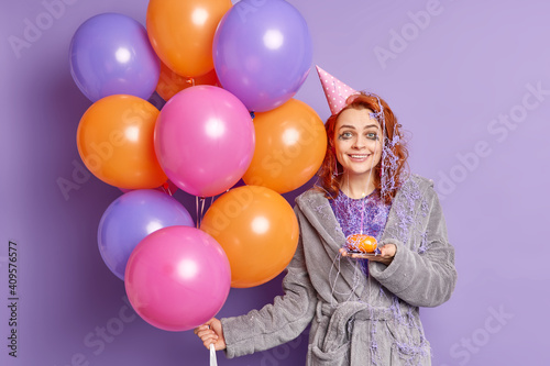 Photo of glad ginger woman smiles toothily dressed in soft dressing gown wears party hat celebrates birthday with friends accepts congratulations isolated over purple background. Celebration concept