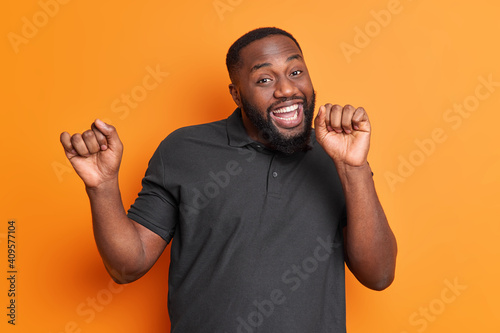 Studio shot of carefree positive black man with thick beard dances carefree has upbeat mood raises arms enjoys favorite music isolated over vivid orange wall. Afro American guy moves actively indoor